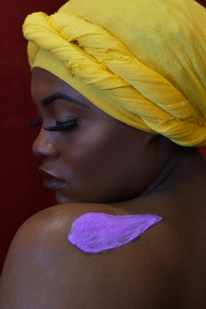 Model with long eyelashes in Bright yellow headwrap with a schmear of our purple colored Calming Seas Body Butter on her shoulder