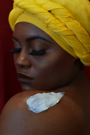 Model with long eyelashes in Bright yellow headwrap with a schmear of our white colored Coconut Shavings Body Butter on her shoulder